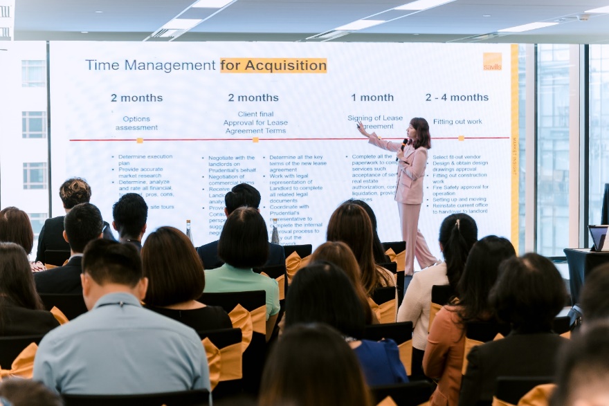 A person standing in front of a large screen with a group of people sitting in chairs  Description automatically generated